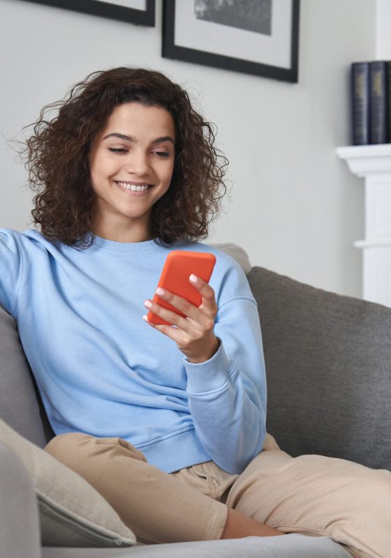 Happy 20s young woman watching social media videos looking at smartphone relaxing on couch, smiling hispanic girl enjoying using online mobile apps doing online shopping on cell phone at home.